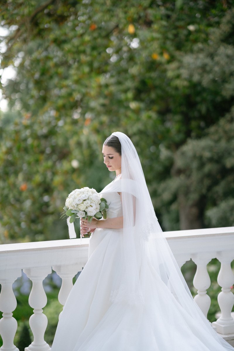 A bride in a white wedding dress holding a bouquet of white roses, standing on a white balcony with greenery in the background.
