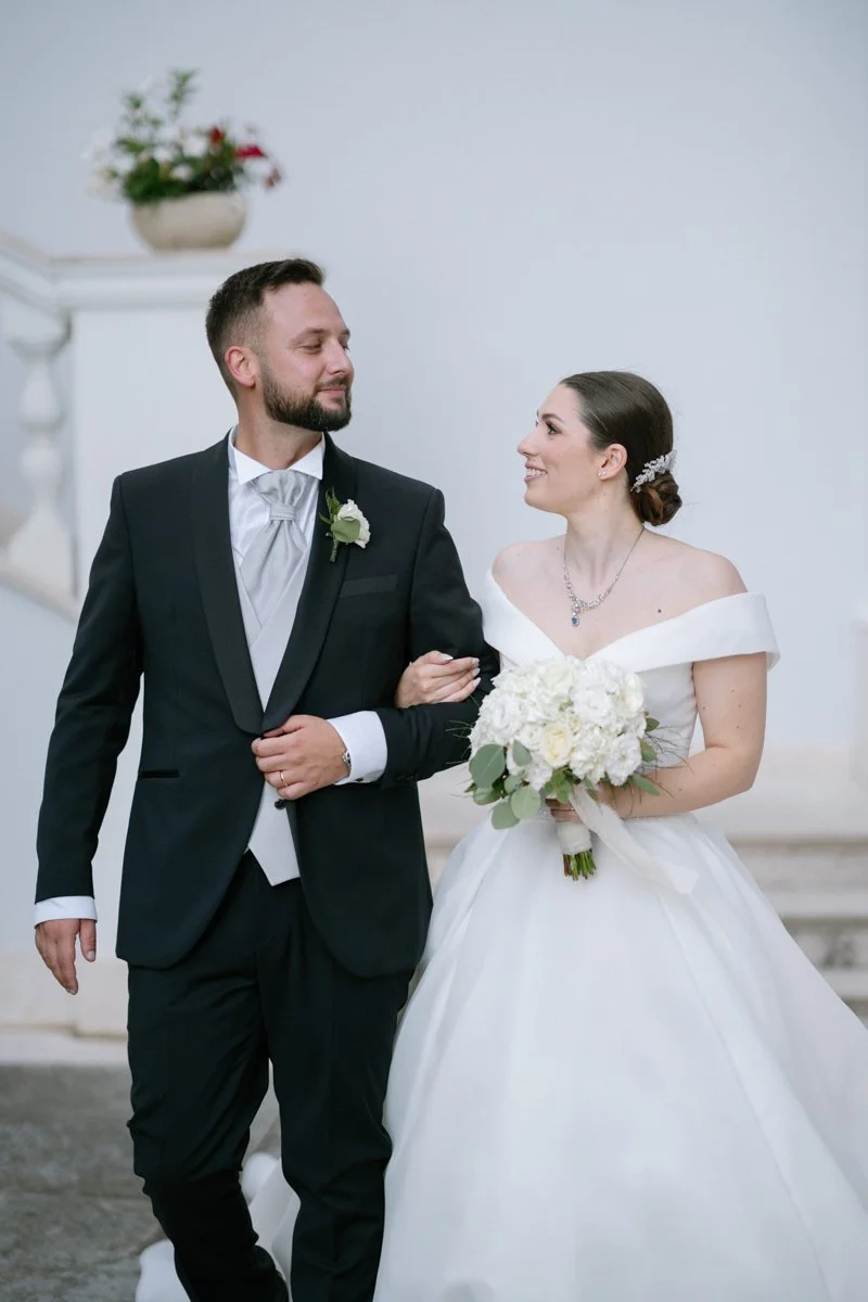 A bride in a white wedding dress and a groom in a black tuxedo walk arm in arm, smiling at each other during their wedding ceremony indoors.