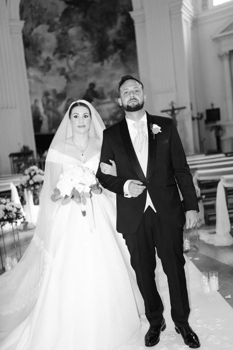 Black and white photo of a bride and groom walking down the aisle inside a church. The bride is wearing a wedding gown and holding a bouquet, and the groom is dressed in a tuxedo with a boutonniere.