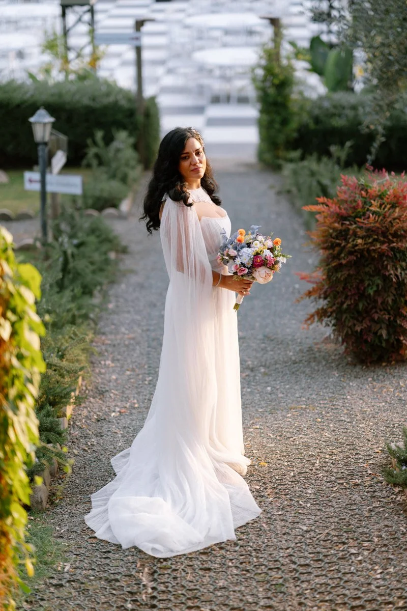 A woman in a white wedding dress holding a bouquet of flowers stands on a garden pathway with greenery on both sides.