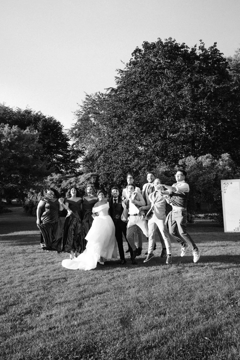 Black and white photo of a group of people standing on grass outdoors, with large trees in the background, celebrating at a wedding.