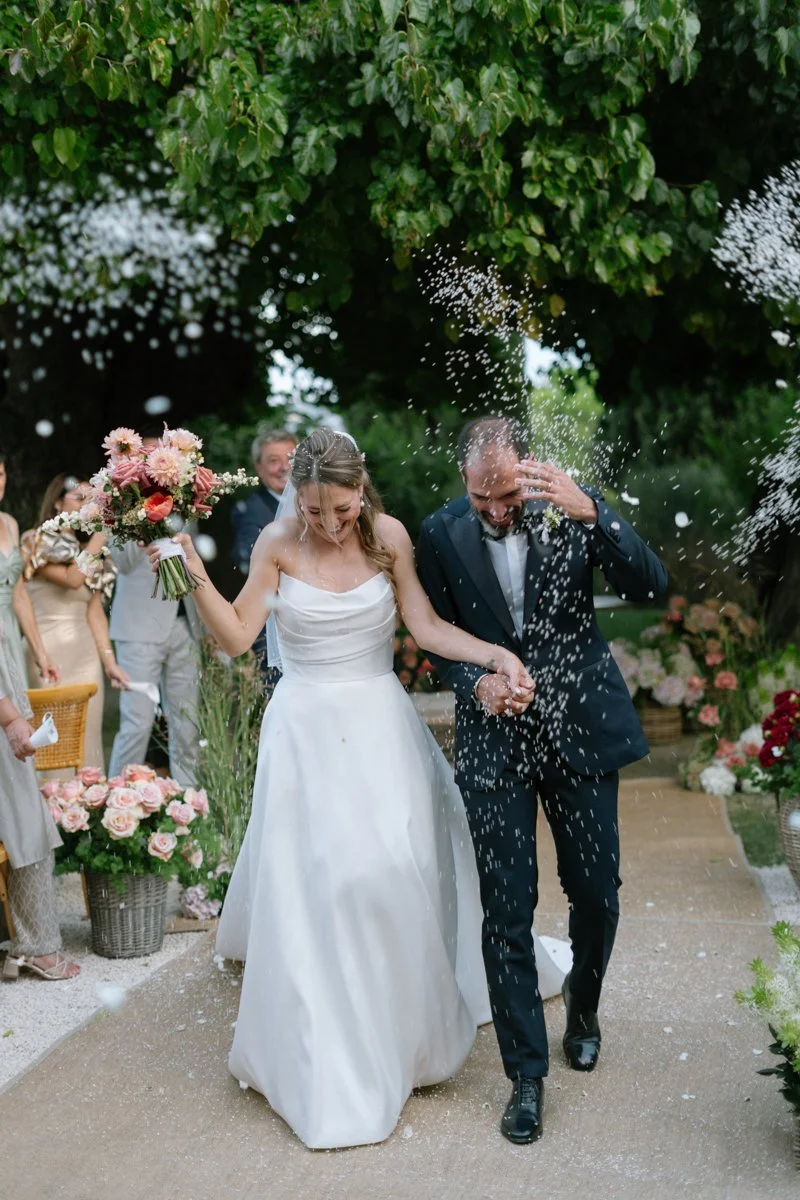 Bride and groom celebrating their wedding outdoors under a tree, with guests around, holding a bouquet and showering the couple with confetti.