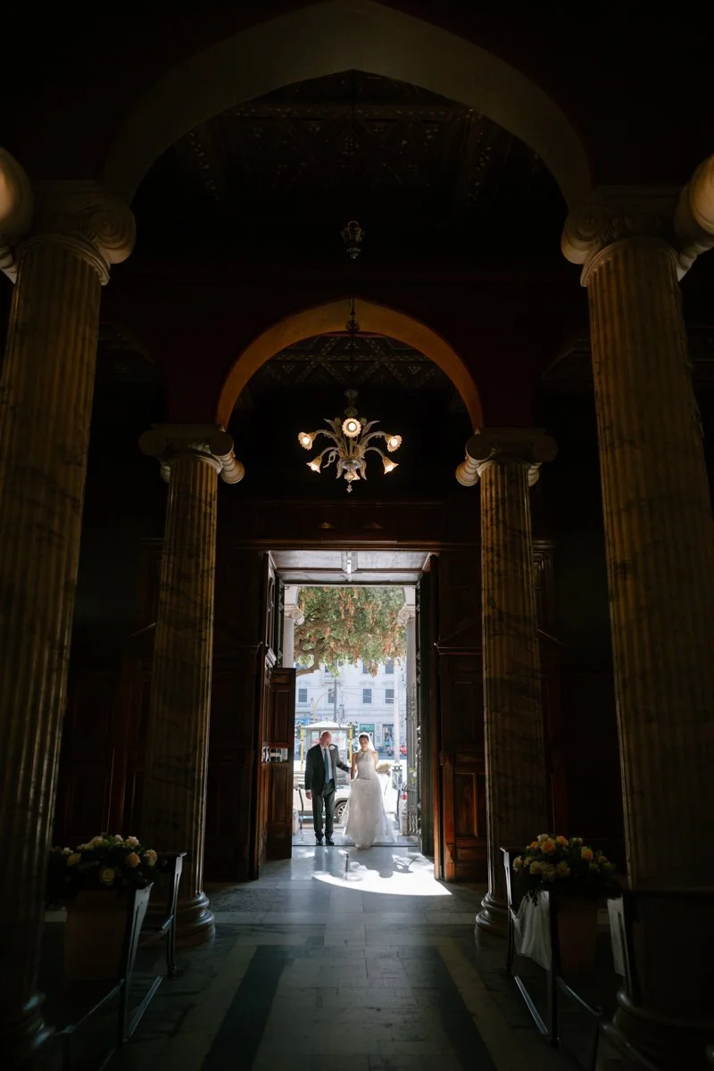 A bride and groom standing in the entrance of a building with tall columns, visible outside through the open doors. Sunlight illuminates the couple and the street outside.