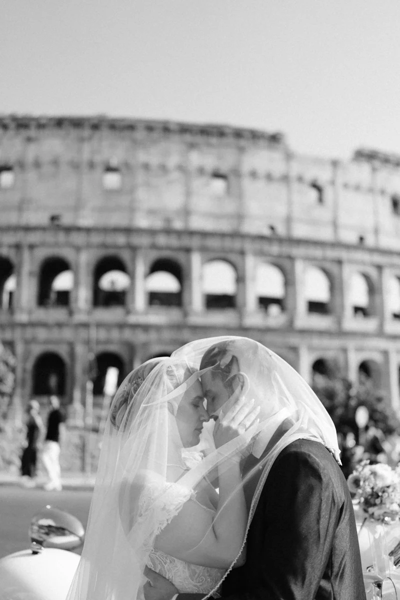 A bride and groom sharing a kiss behind a wedding veil with the Roman Colosseum in the background.