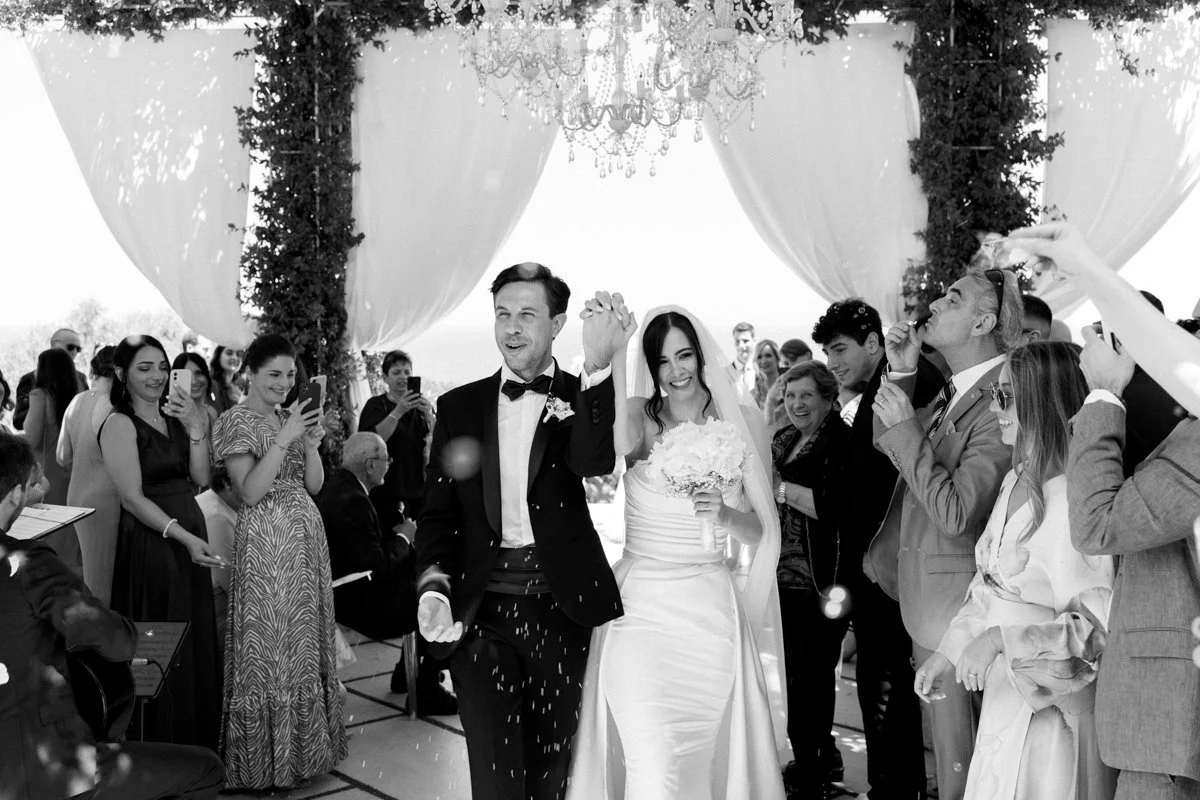 Black and white photo of a wedding celebration with a bride and groom walking under a decorated arch, surrounded by guests taking photos and smiling.