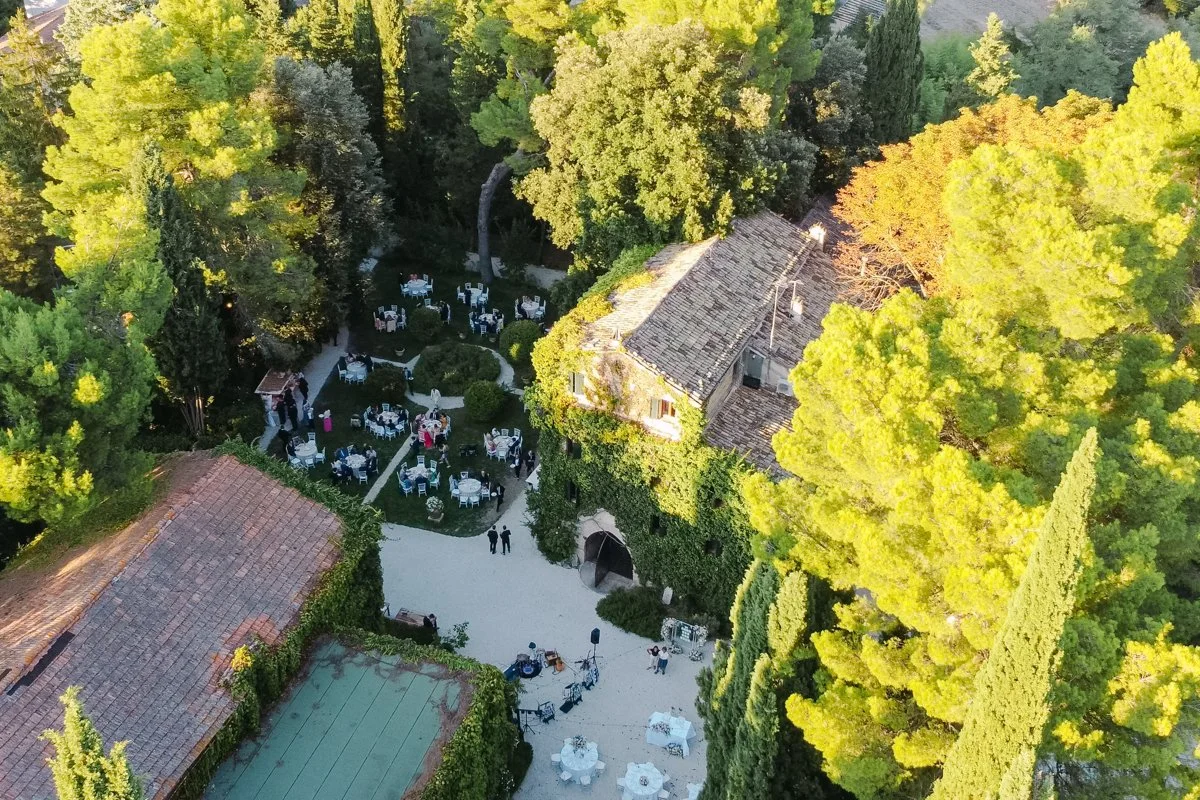 An aerial view of an outdoor event with tables and chairs set up for a gathering, surrounded by lush green trees and a stone building with a tiled roof.