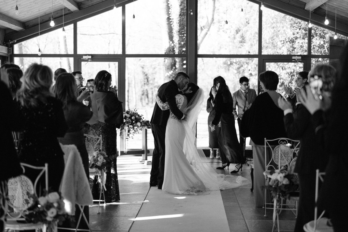 Black and white photo of a wedding ceremony with a bride and groom sharing a kiss in the center of a decorated venue, surrounded by guests clapping and taking photos.