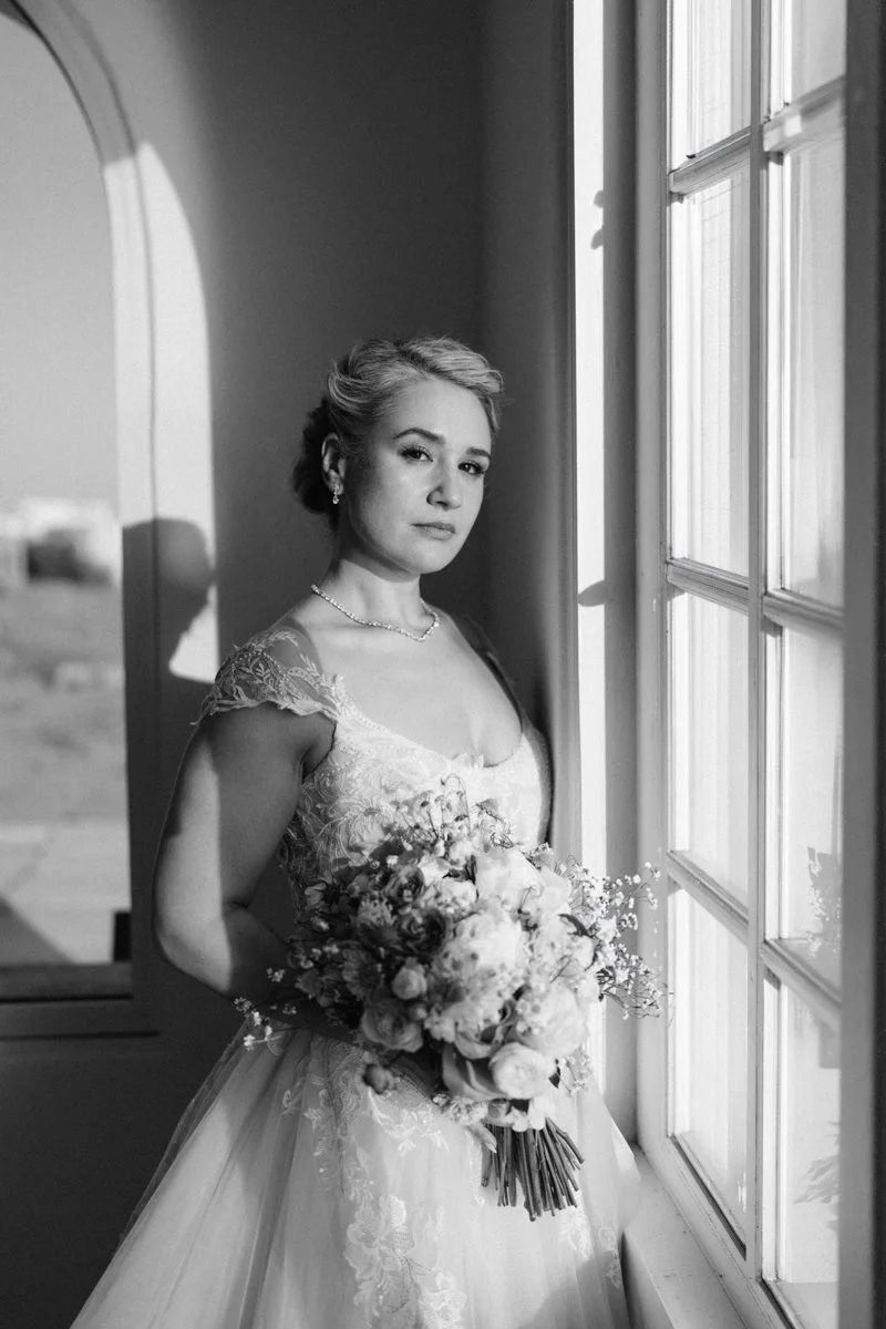A woman in a wedding dress holding a bouquet, standing by a window in a room with sunlight.