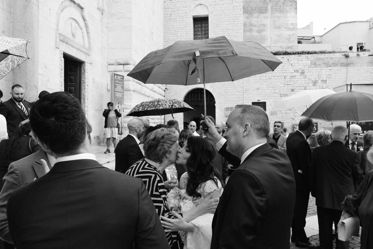 A black-and-white photo of a wedding scene outside a historic building. Several people in formal attire hold umbrellas during rain. A woman in a wedding dress is kissing an older woman, possibly her mother. A man holding an umbrella over the bride is