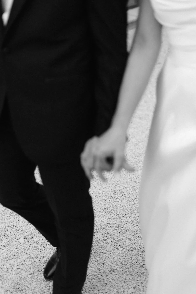 Close-up of two people holding hands, one wearing a black suit and the other a white dress, on a textured surface.