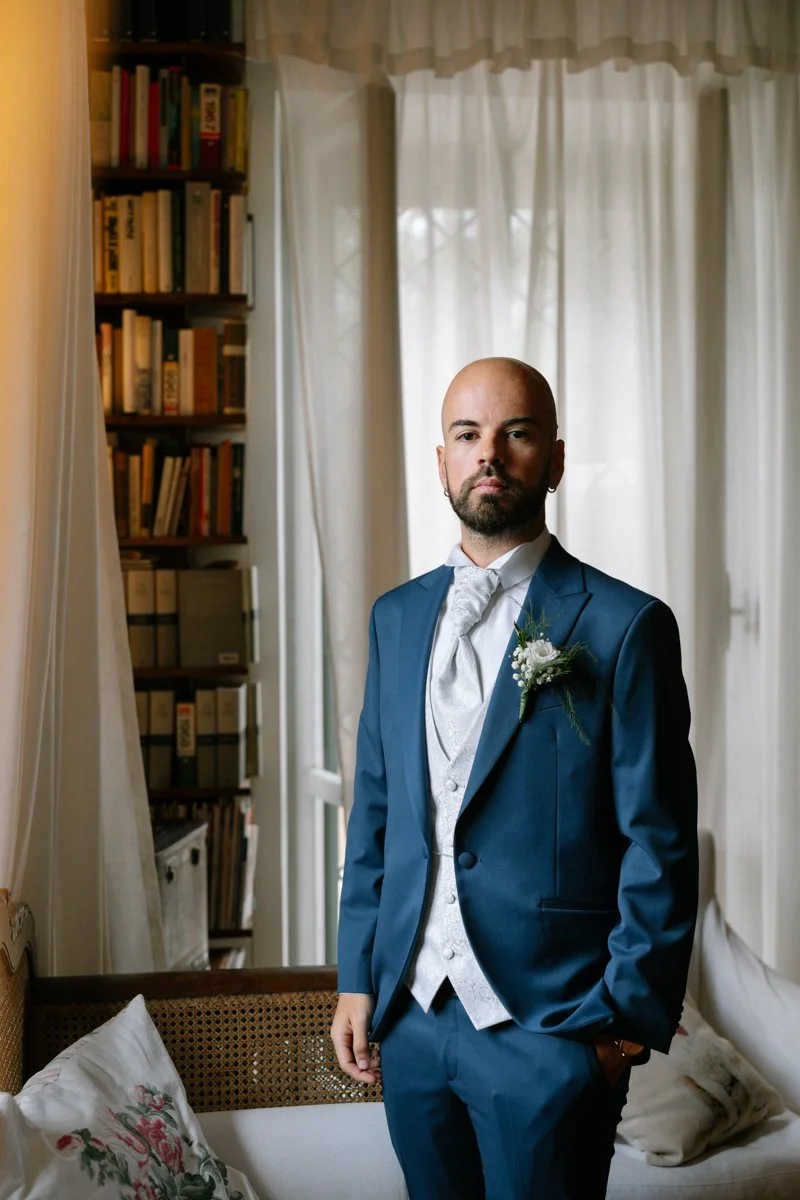 A man dressed in a blue suit with a boutonniere standing in a cozy room with bookshelves and white curtains.