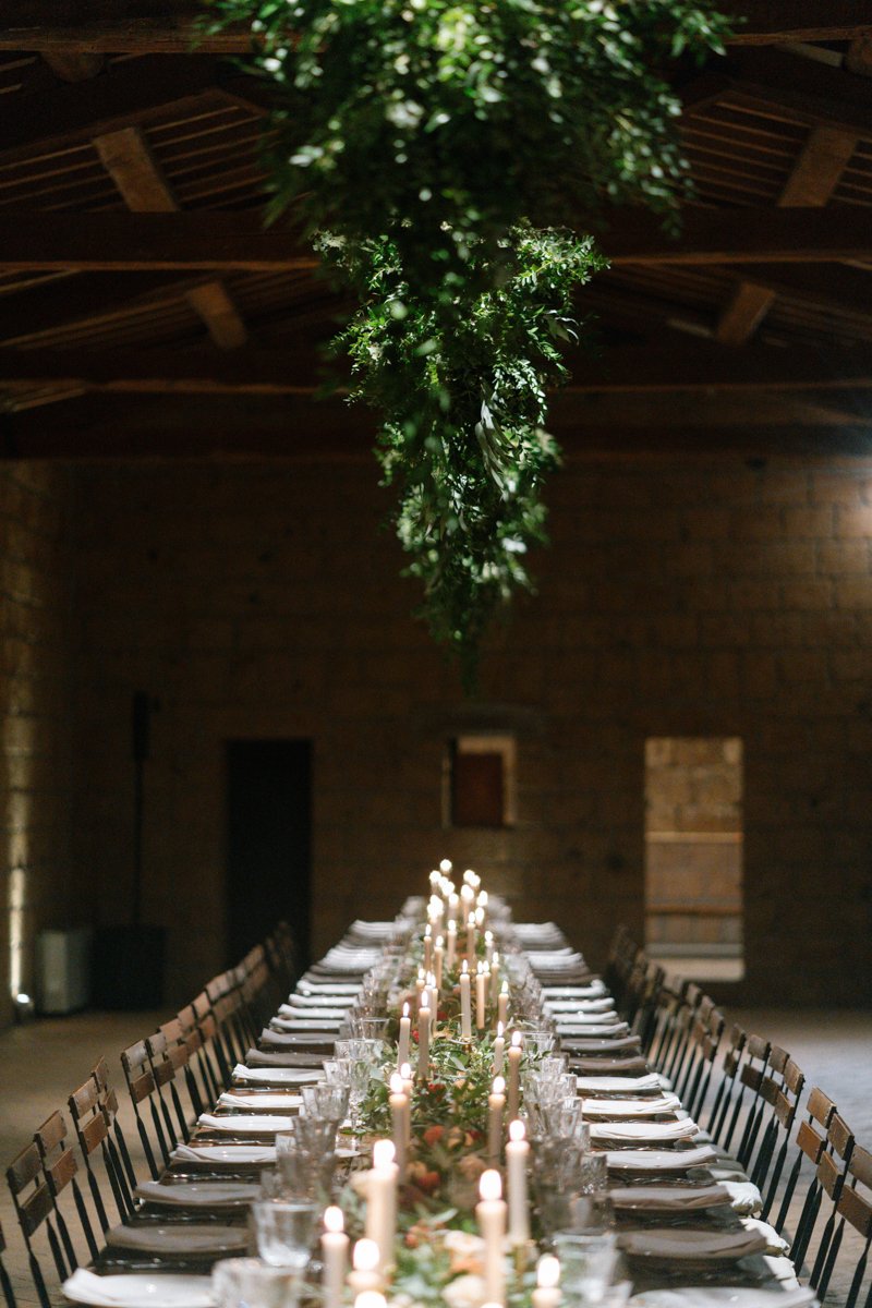 Long dining table set for a formal event with multiple lit candles, glassware, and folded napkins, decorated with greenery and flowers, in a rustic wooden venue with a hanging green floral centerpiece above.