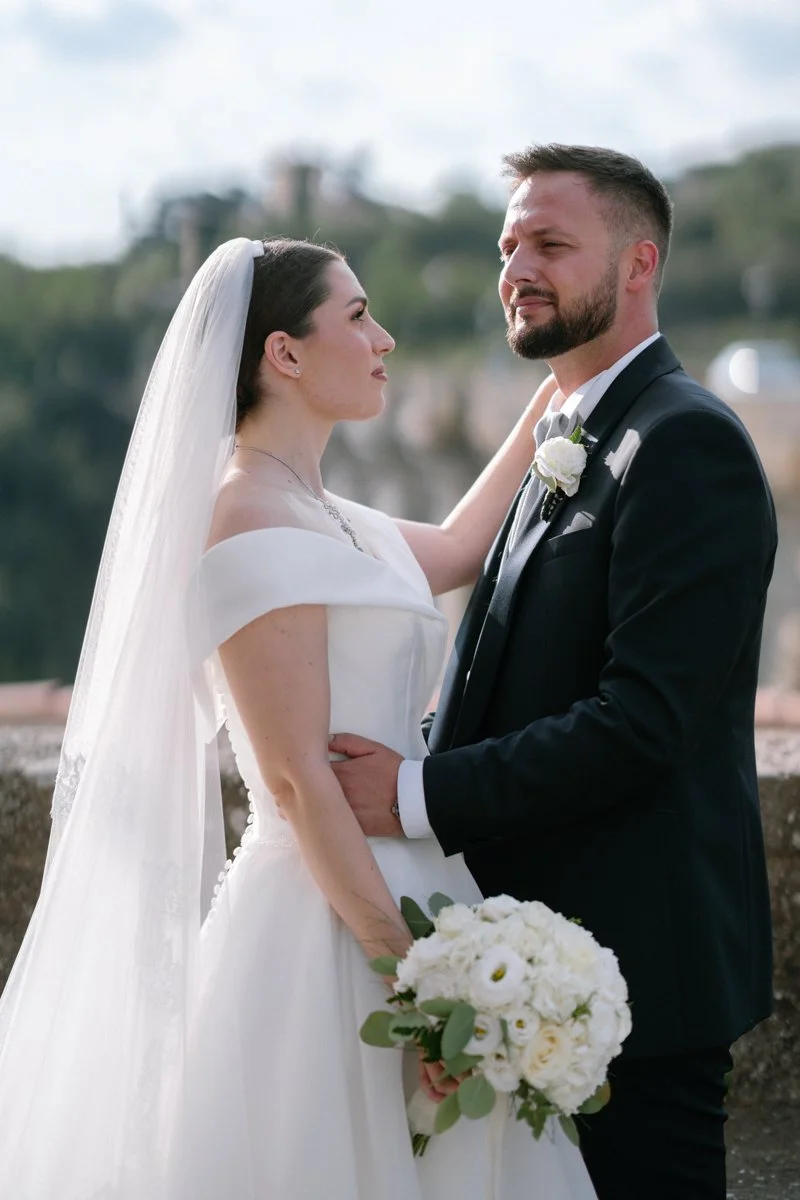 A bride and groom stand close together outdoors, gazing into each other's eyes, with the bride holding a bouquet of white flowers.