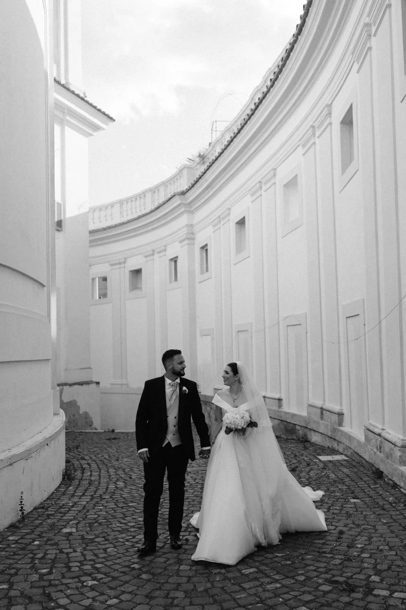 A black and white photograph of a bride and groom walking hand in hand in a curved courtyard with white walls and cobblestone ground. The bride is holding a bouquet and wearing a wedding dress with a veil, while the groom is in a suit with a boutonni