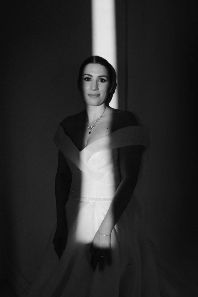 A woman in an elegant dress, standing in front of a dark background, illuminated by a narrow beam of light, in black and white photography.