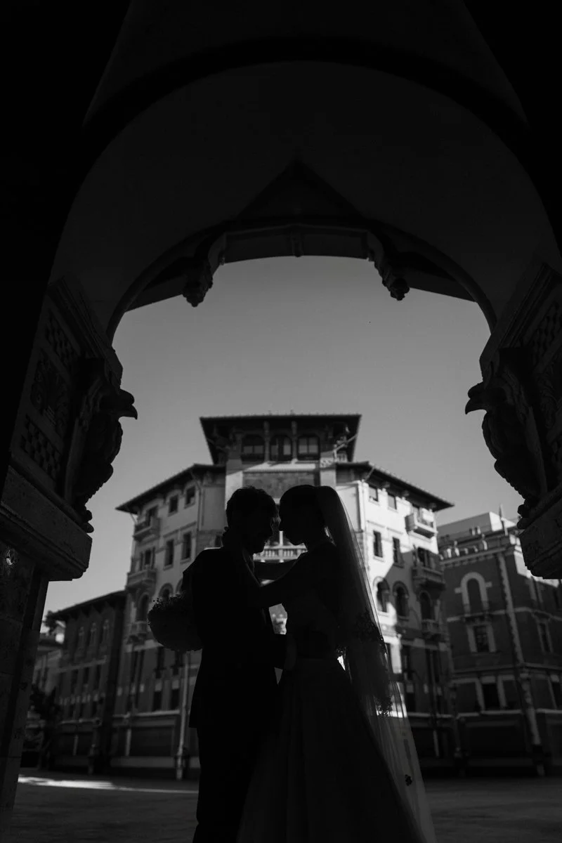 Silhouettes of a bride and groom standing close together under an ornate archway in front of a large, historic building during sunset.