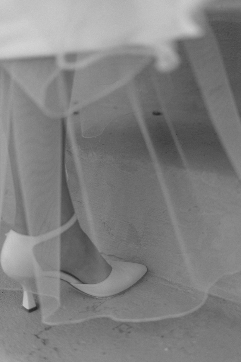 Close-up of a woman’s leg and white high heel shoe, with a sheer dress. The photo is in black and white.