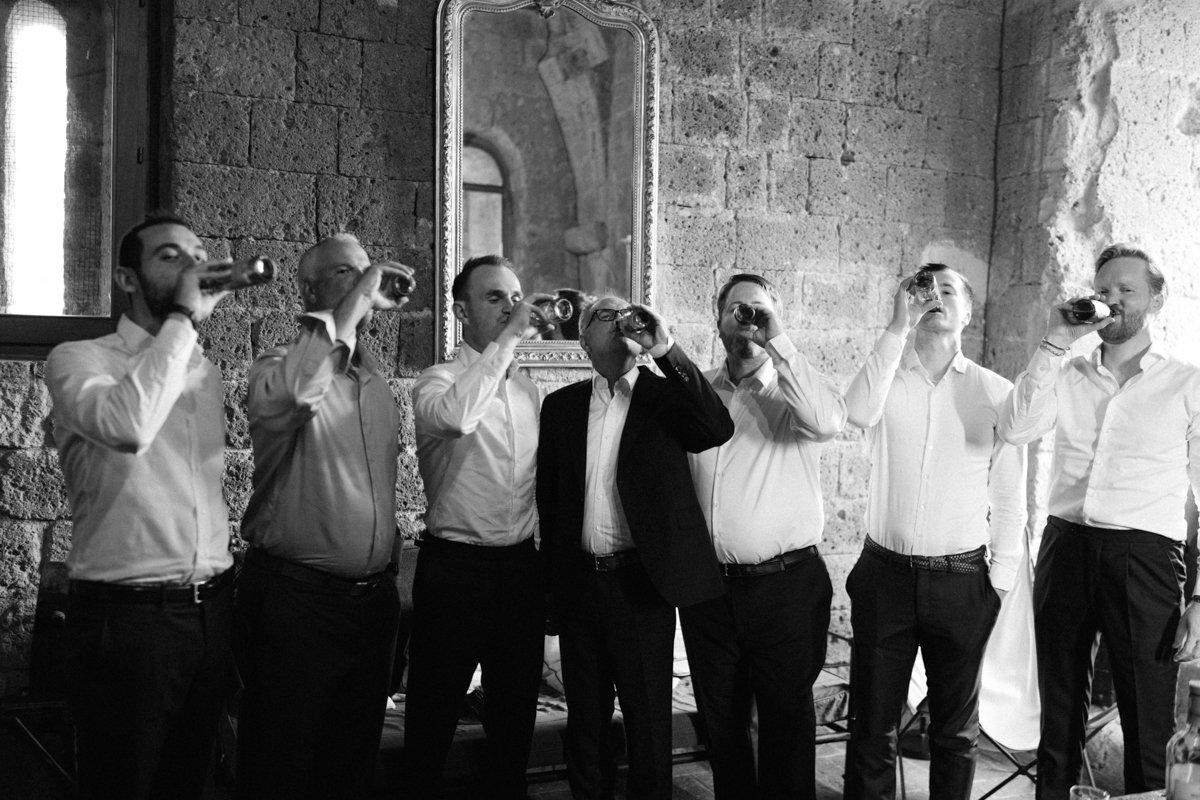 Seven people in formal attire drinking beer in an indoor setting with stone walls and a mirror.