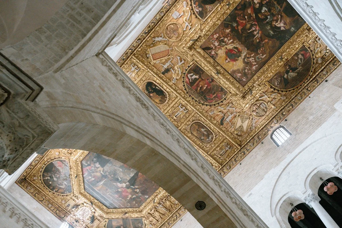 View of an ornate, gold-framed ceiling with religious paintings inside a church or cathedral, viewed from below near an archway.