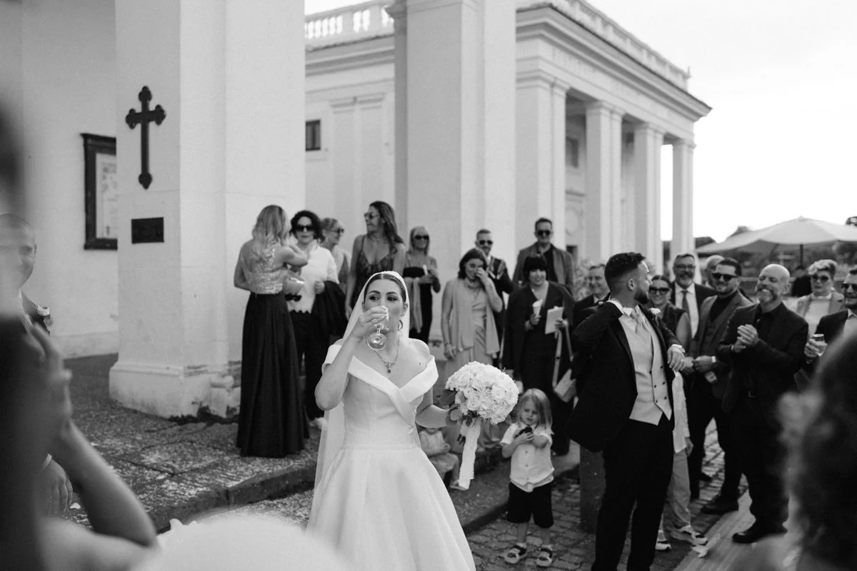 Bride in wedding dress holding a bouquet and drinking champagne, surrounded by wedding guests outdoors in front of a church or chapel.
