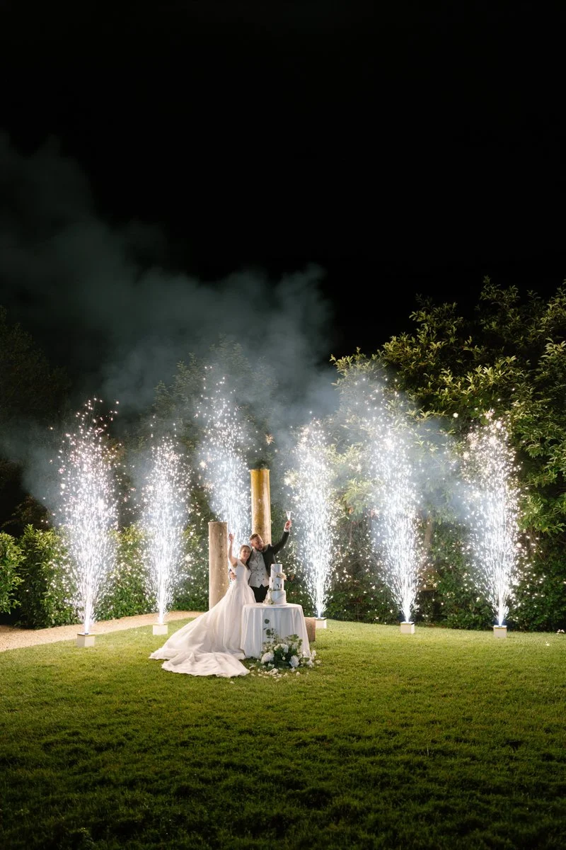 Wedding celebration with fireworks and sparklers at night, featuring a bride and groom standing behind a table with a wedding cake on it, surrounded by greenery.