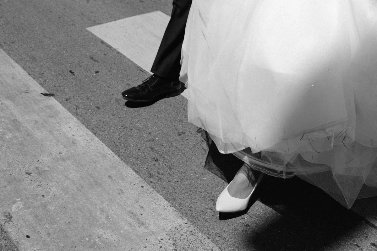 Close-up of a bride in a wedding dress with white heels and a groom in black shoes and pants standing on a crosswalk.