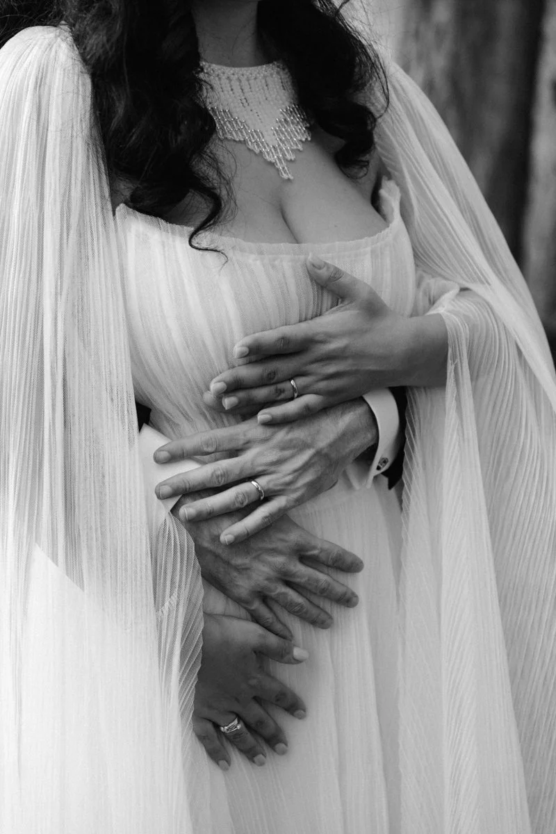 A woman in a wedding dress, with her hands and another person's hands on her waist and stomach, showing rings on her fingers, in black and white.