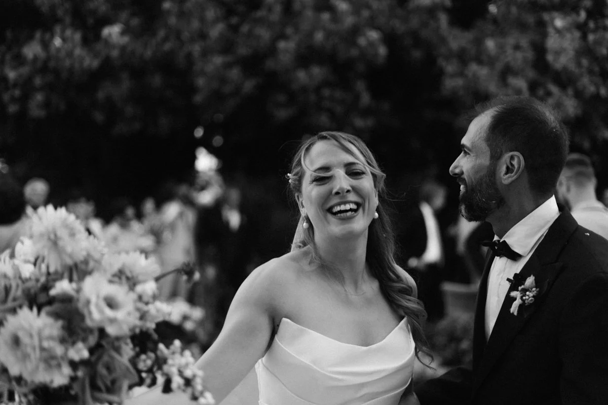 A bride and groom celebrating at their wedding reception, with the bride laughing and the groom smiling at her, surrounded by wedding guests and flowers.