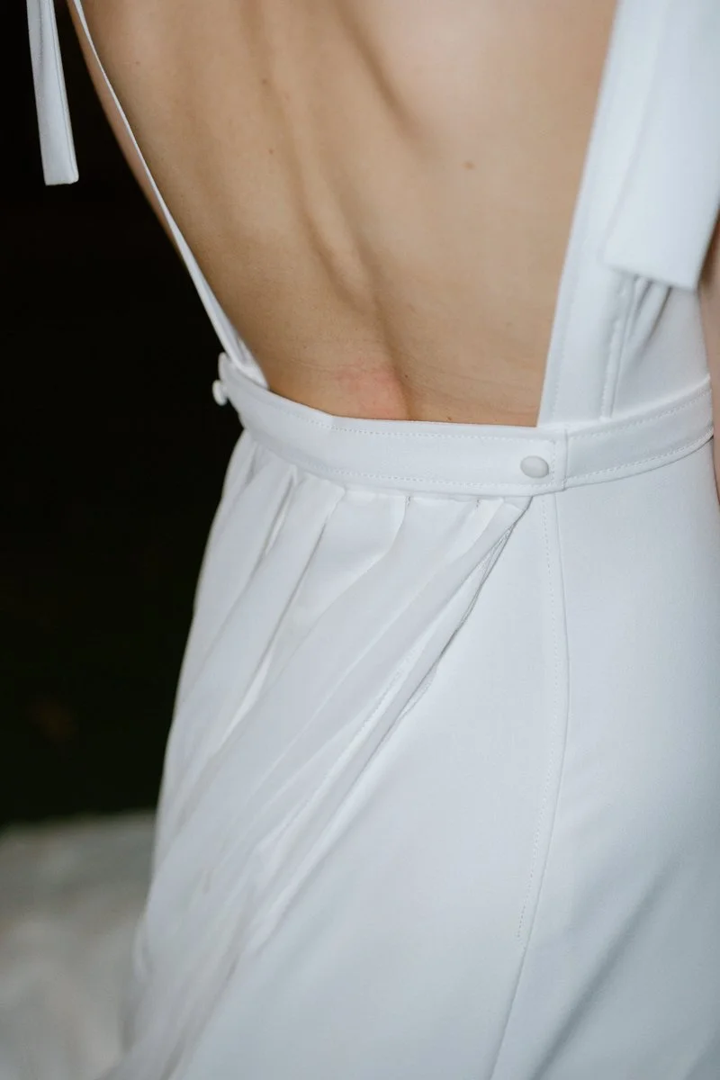 Close-up of a person wearing a white, backless dress with pleats, showing part of their bare back and side.