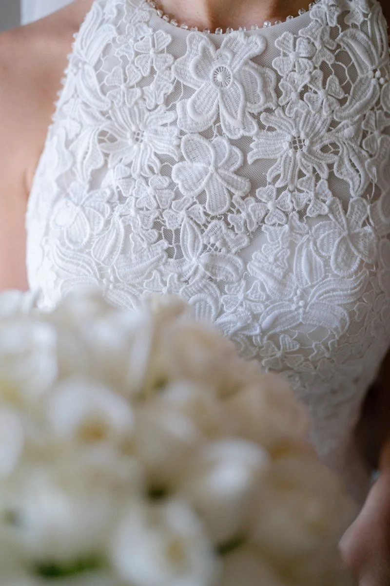 Close-up of a woman wearing a white lace wedding dress with a floral pattern, holding a bouquet of white flowers.