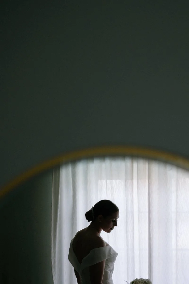 A woman in an off-shoulder dress standing near a window with sheer curtains, viewed in a mirror.