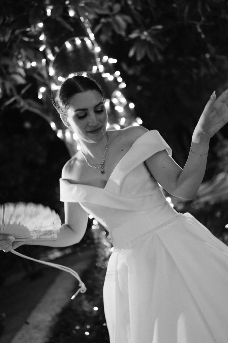 A woman in an off-shoulder white dress holding a fan with fairy lights and trees in the background.