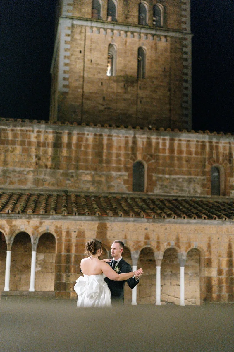 A bride and groom dancing outdoors at night in front of a historic brick building with arches and a tower.