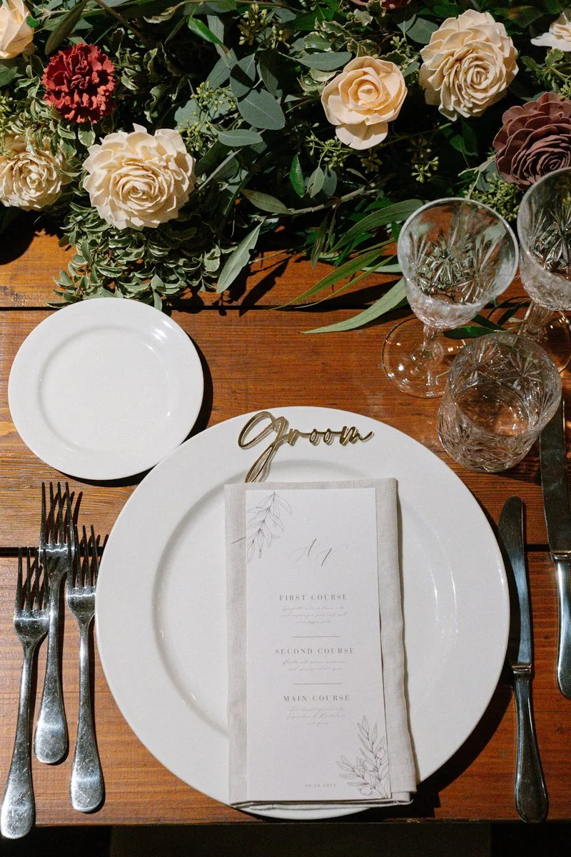 A table setting with a white plate, cloth napkin, and a menu card, surrounded by wine glasses and a floral arrangement with roses and greenery on a wooden table.