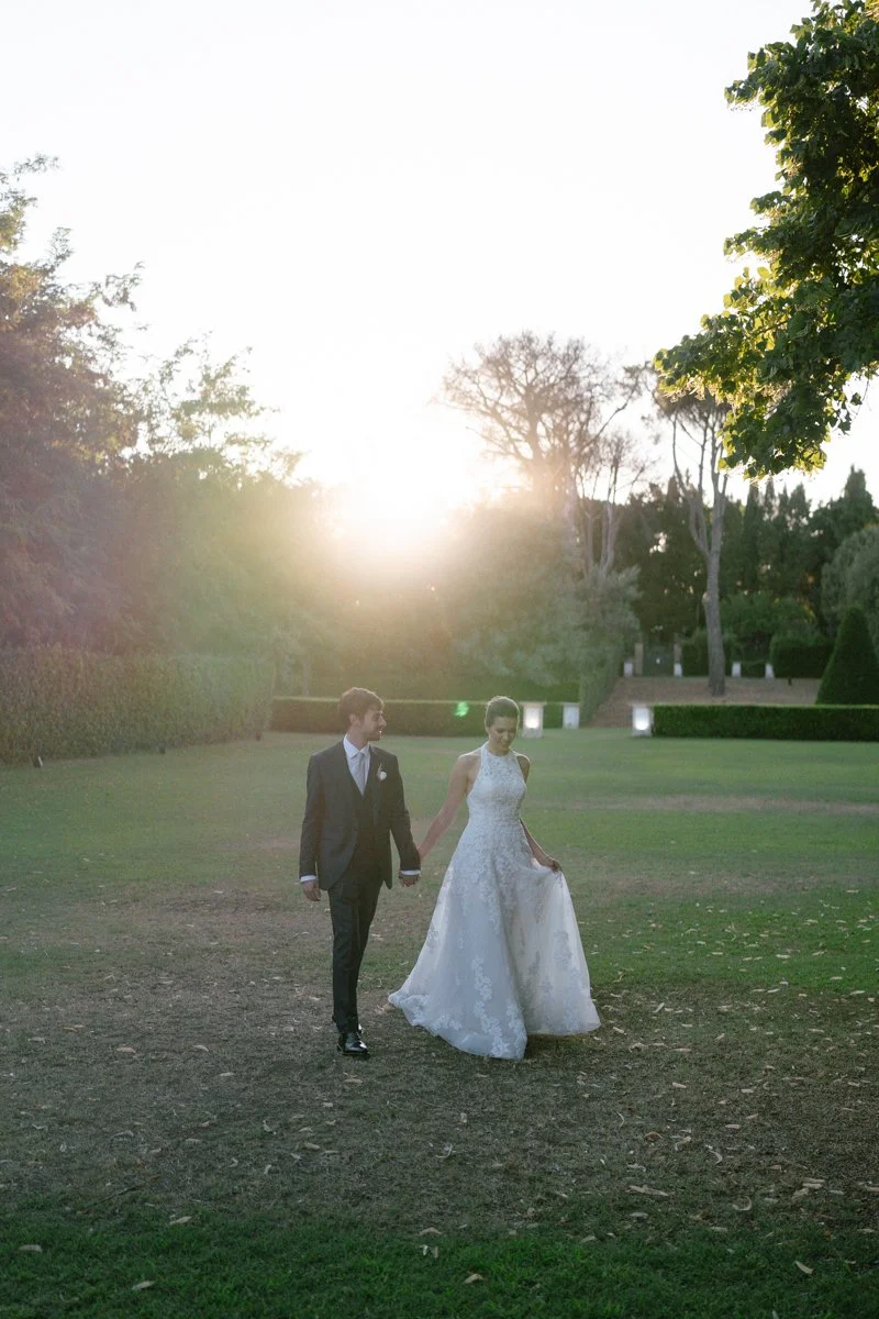 A bride and groom walking hand in hand outdoors on a grassy area during sunset, with trees and bushes in the background.