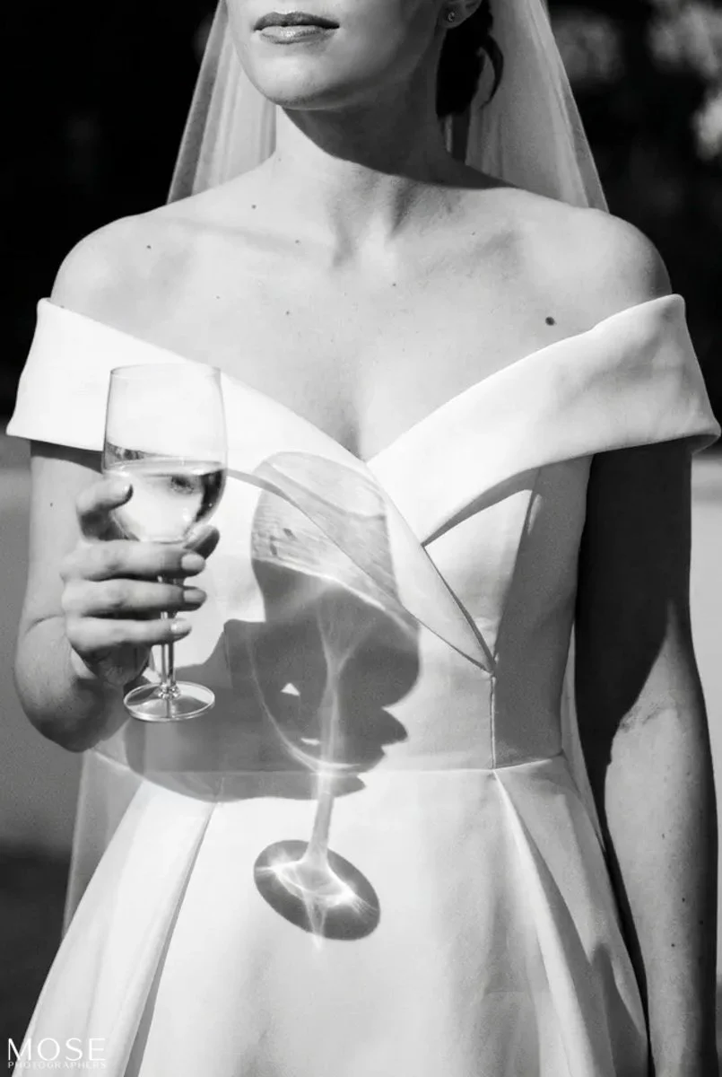 Black and white photo of a woman in an off-shoulder dress holding a glass of wine, with sunlight casting reflections and shadows on the glass and dress.