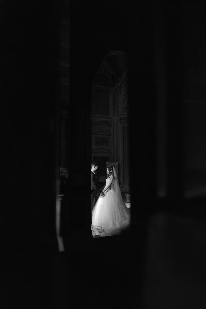 A black and white photo of a bride and groom holding hands inside a church or cathedral, viewed through a narrow opening or gap in the wall or door.
