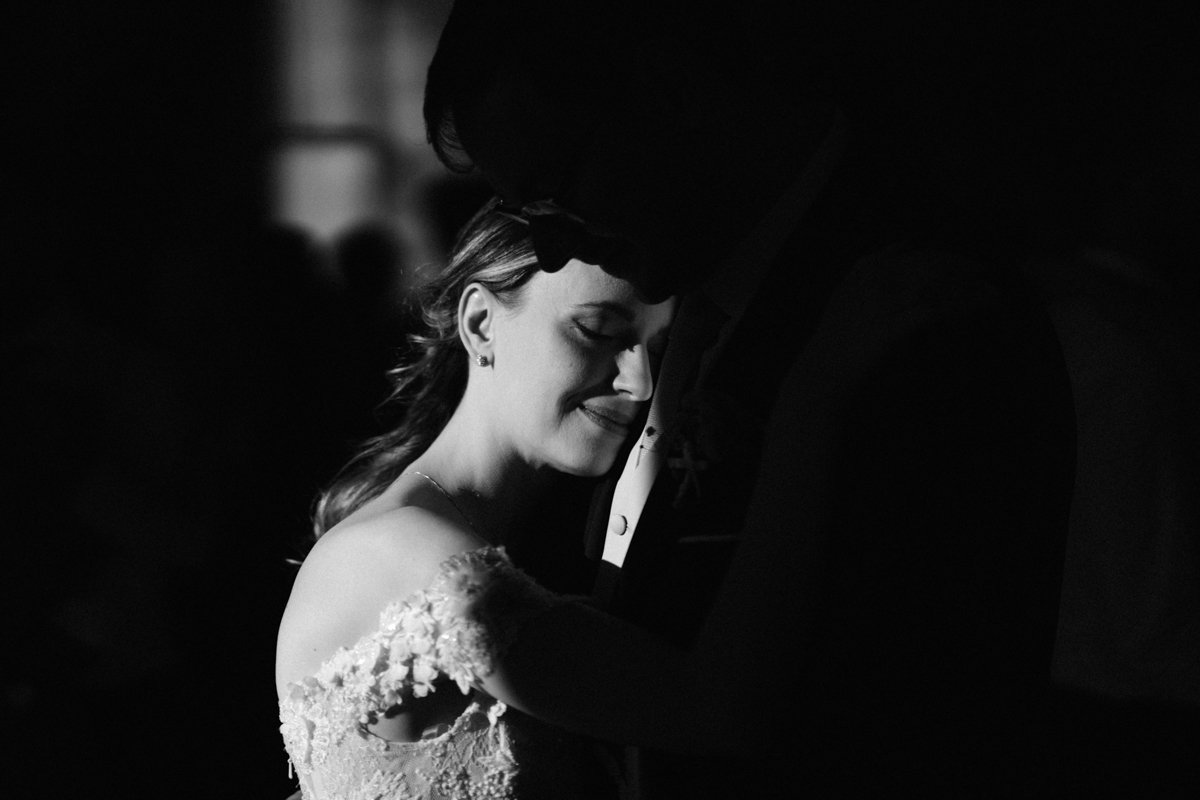 Black and white photo of a woman in a wedding dress smiling with her eyes closed, leaning into a man in a suit, who is positioned close to her.
