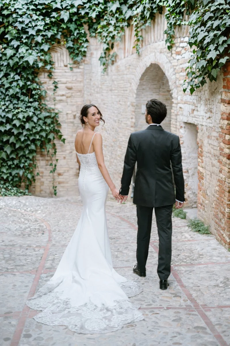 A bride and groom walking hand in hand on a cobblestone path, with the bride smiling and looking back at the camera, surrounded by stone and brick walls covered partially in green ivy.
