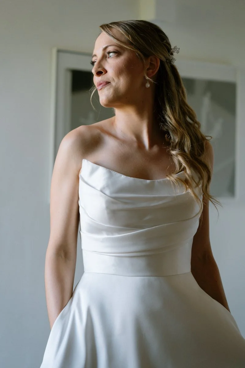 A bride in a white satin wedding dress with a Strapless neckline, standing indoors, looking to her left.