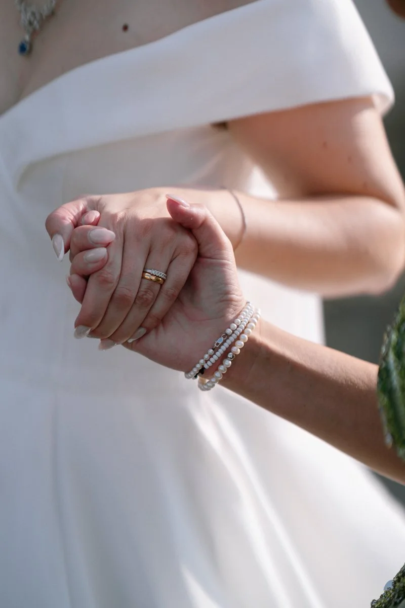 Close-up of two women holding hands, one wearing a wedding ring and pearl bracelets, the other wearing a white dress, possibly at a wedding or special event.