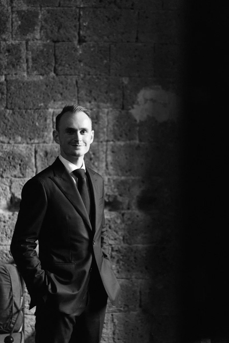 A young man in a suit standing against a brick wall, smiling with his hands in his pockets.