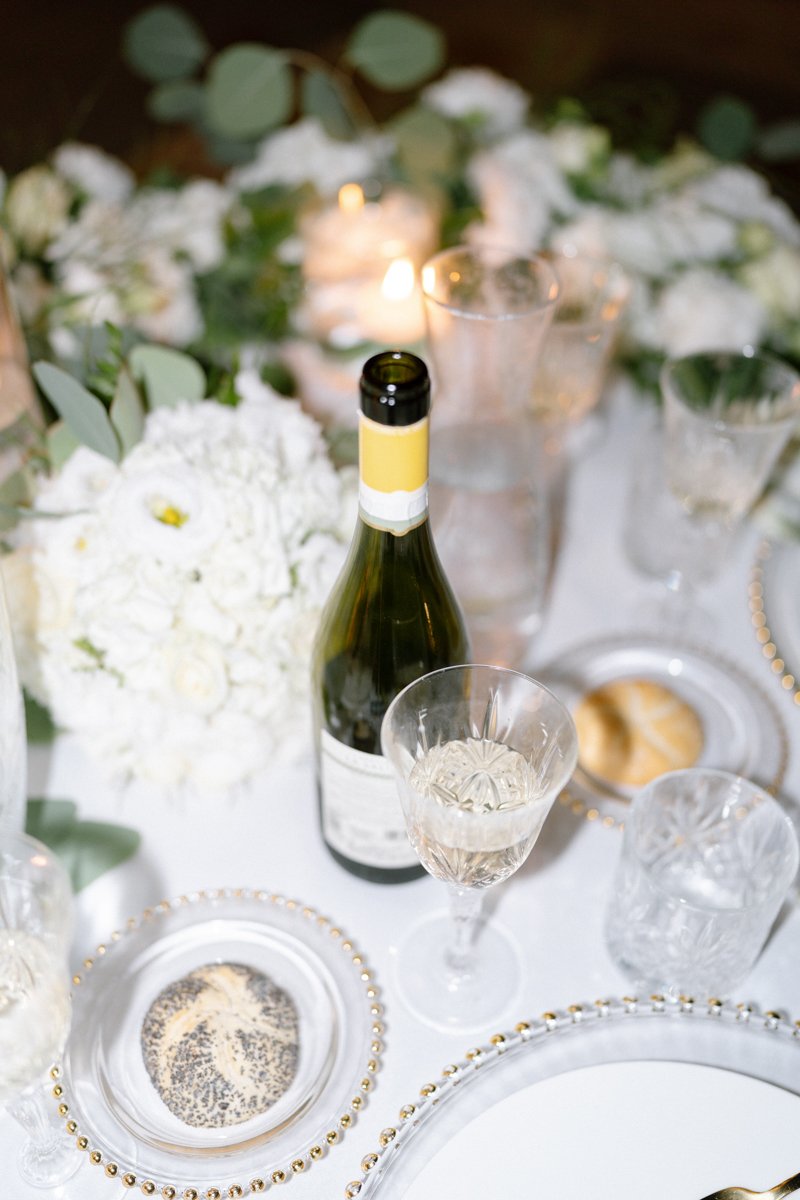 A table set for a celebration with a bottle of white wine, empty glasses, a floral centerpiece of white flowers and greenery, and candles.