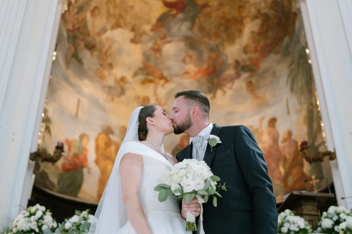 A bride and groom sharing a kiss during their wedding ceremony inside a church with a mural in the background and floral arrangements in the foreground.