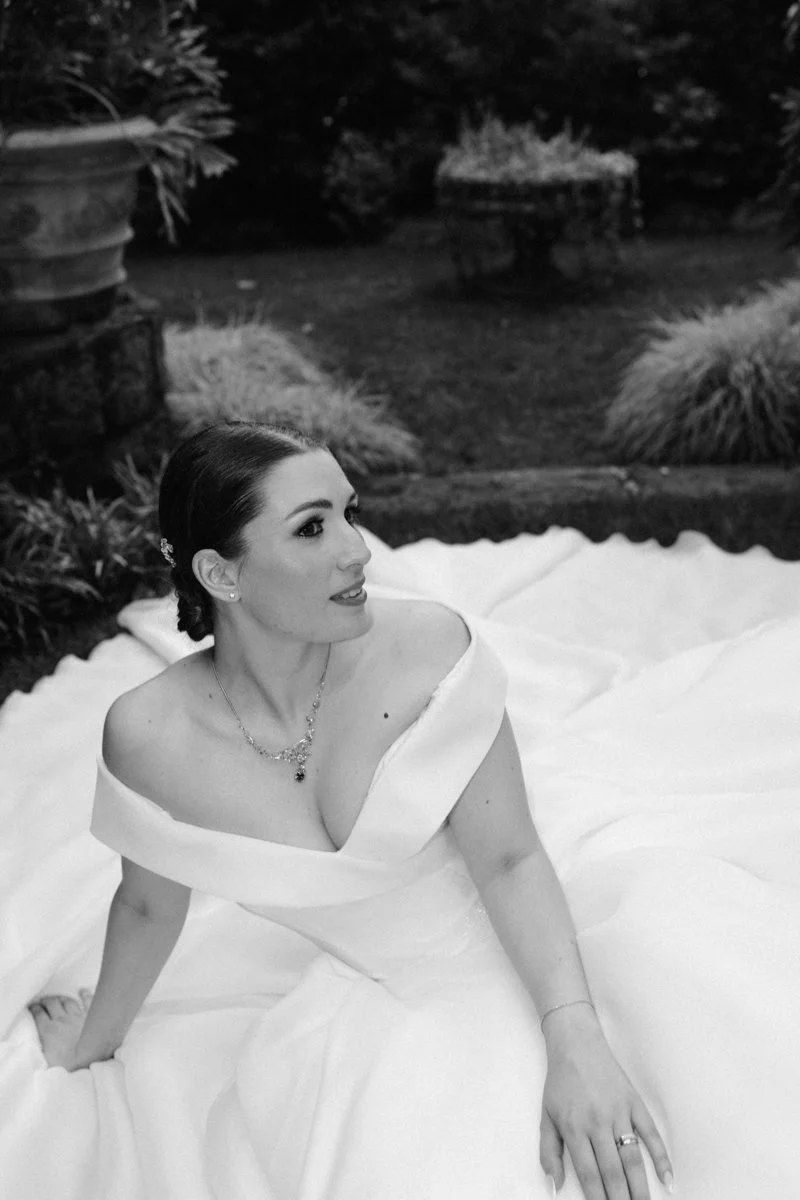 A woman in a wedding dress sitting outdoors on a blanket in a garden, looking to her right, with landscaping and plants in the background.