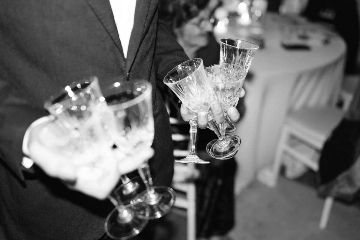 A person in formal attire holding several empty wine glasses at a gathering or celebration.