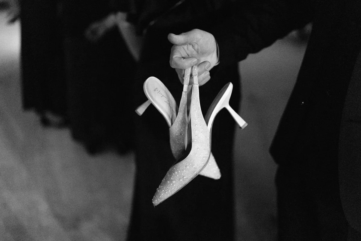 A person holding a pair of high-heeled shoes with pointed toes and high heels, held by the straps in their hand.