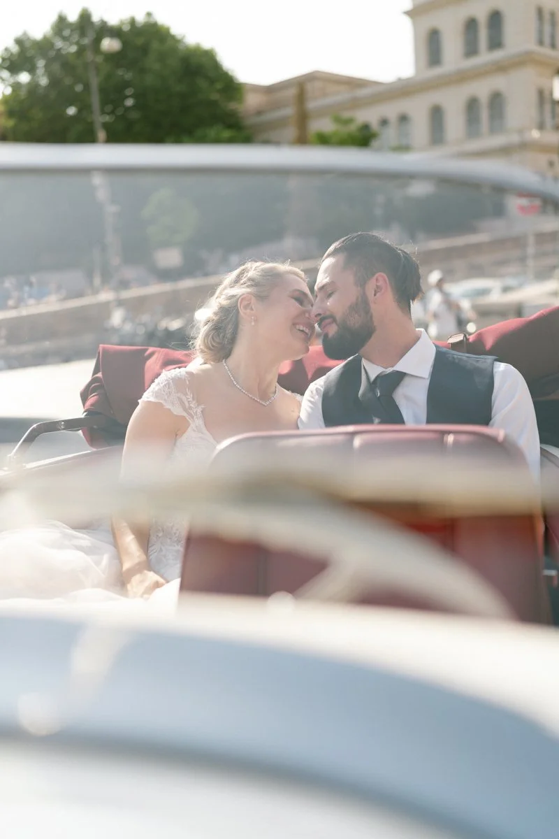 A newly married couple sharing a kiss and smiling in a vintage car during daytime, with a cityscape and river in the background.