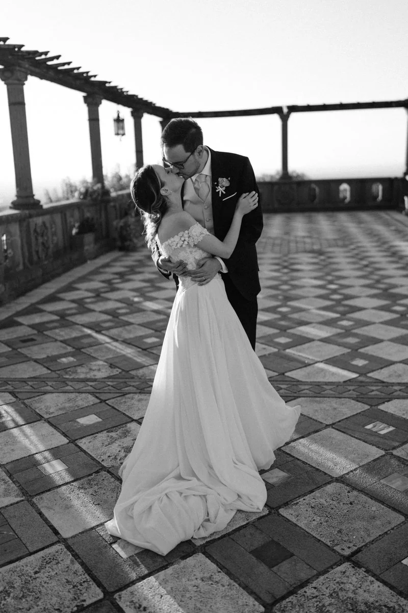 Black and white photo of a bride and groom sharing a kiss on a tiled outdoor terrace with a pergola structure in the background.