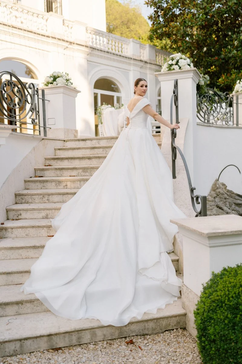 A bride in a white wedding dress walking up marble stairs outside a white building with ornate railings and greenery.
